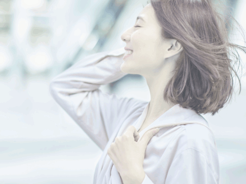 風になびく髪を揺らしながら、右手を耳に添えてほほ笑む横向きの女性の写真。自信に満ちた表情が印象的。